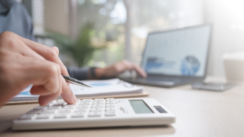 Osoba računa troškove na digitronu uz dokumentaciju i otvoren laptop