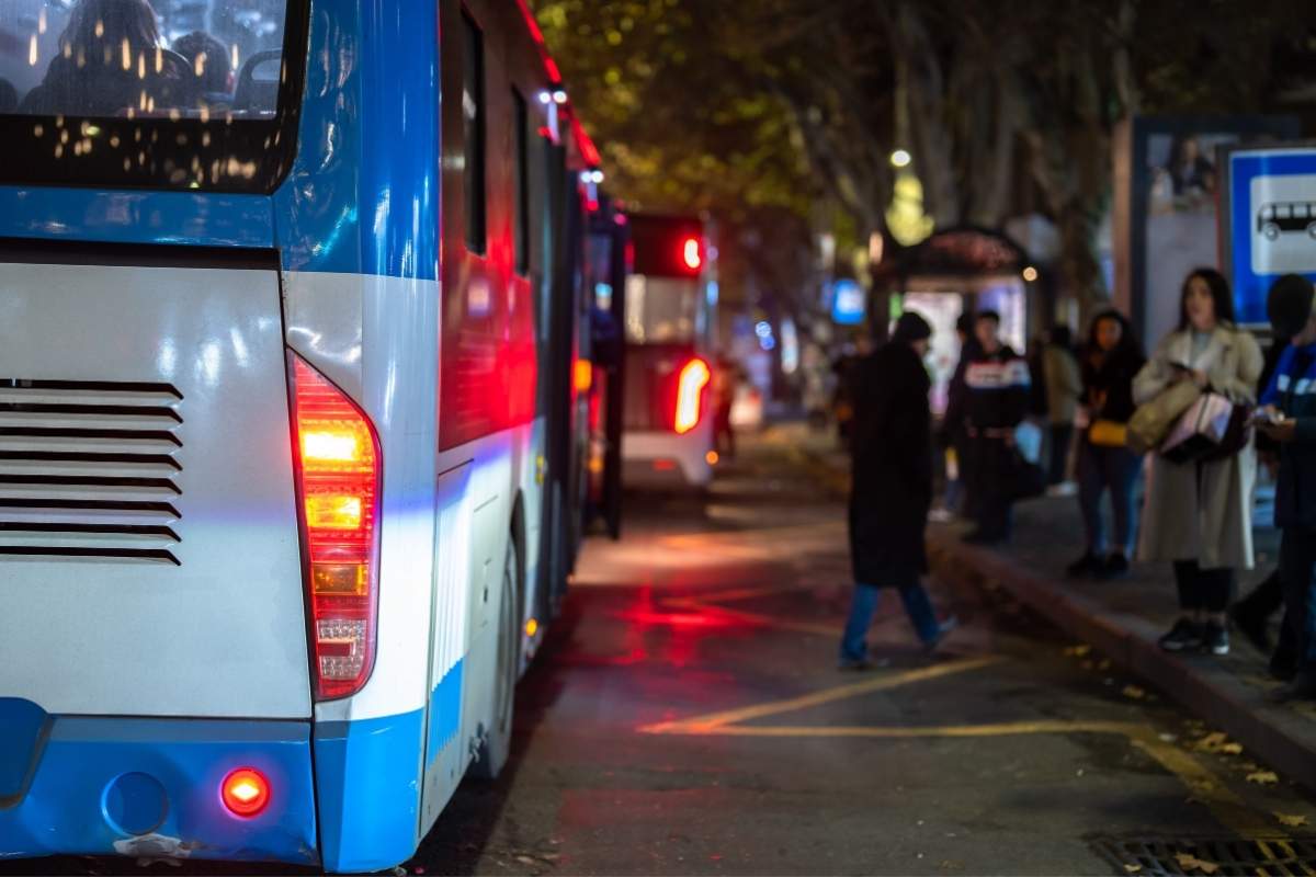 osobe čekaju autobus na autobuskom stajalištu, ispred njih 2 autobusa
